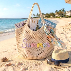 K&ouml;nnte beinhalten: Beige geh&auml;kelte Tragetasche mit dem Wort "Summer" in Pastellfarben. Die Tasche ist mit Zeitschriften gef&uuml;llt und ein floraler Schal ist am Griff befestigt. Ein Strohhut mit marineblauem Band und Sonnenbrille liegen im Sand.