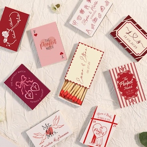 Könnte beinhalten: Eine Sammlung personalisierter Streichholzschachteln in verschiedenen Designs. Einige Schachteln zeigen Namen, Daten und den Satz „The Perfect Match“. Farben sind Rot, Rosa und Weiß, mit dekorativen Elementen wie Herzen, Blumen und Bändern. Ein Schachtel zeigt die Streichhölzer.
