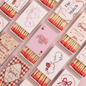 May include: A collection of matchboxes with red-tipped matches. The boxes are decorated with diverse designs, including hearts, floral patterns, and text like "The Perfect Match" and names with dates. The color scheme incorporates pink, red, and white.