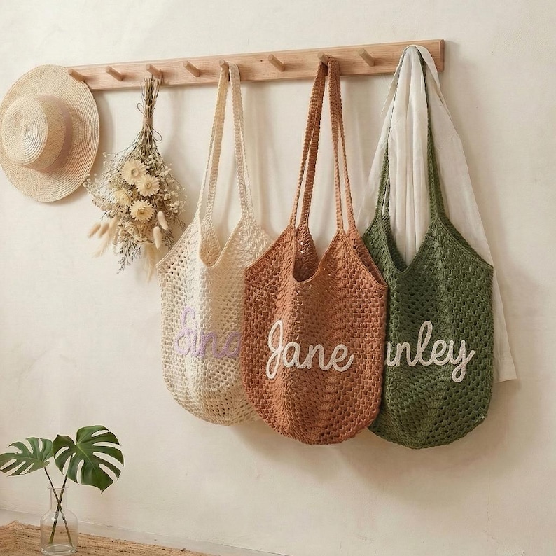 K&ouml;nnte beinhalten: Vier geh&auml;kelte Tragetaschen in Creme, Braun und Gr&uuml;n h&auml;ngen an einer Holzgarderobe. Die cremefarbene Tasche hat den Namen "Sine" in Lila gestickt, die braune Tasche "Jane" in Wei&szlig; und die gr&uuml;ne Tasche "inley" in Wei&szlig;. Ein Strohhut und Trockenblumen sind ebenfalls an der Garderobe.