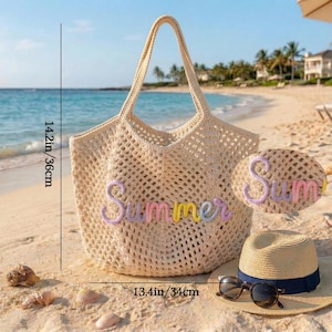 K&ouml;nnte beinhalten: Beige H&auml;keltasche mit dem Wort "Summer" in Pastellfarben. Die Tasche ist 36 cm hoch und 34 cm breit. Ein Strohhut mit marineblauem Band und Sonnenbrille liegen neben der Tasche am Strand.