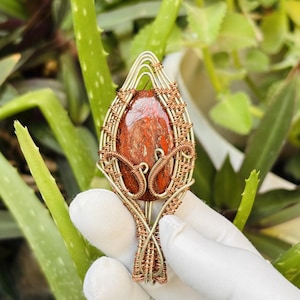 May include: A handcrafted pendant featuring a red and brown stone encased in a wire-wrapped design. The pendant has a teardrop shape, with intricate wire detailing in gold and copper tones. The background is blurred with green foliage.
