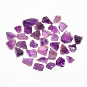 Puede incluir: Un conjunto de cristales de amatista en bruto, en tonos de púrpura variados, desde lavanda claro hasta violeta oscuro. Los cristales, de formas irregulares, se presentan sobre un fondo blanco, mostrando su textura natural.