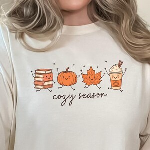 Op de afbeelding: Een ivoorkleurig t-shirt met een grafische print van een stapel boeken, een pompoen, een esdoornblad en een koffiebeker, allemaal met lachende gezichten en ledematen. De tekst "cozy season" staat in een zwart cursief lettertype onder de figuren.