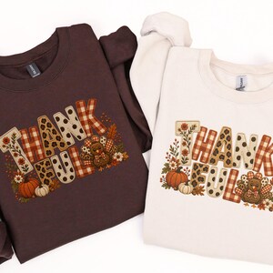Op de afbeelding: Een donker chocoladebruin sweatshirt en een zandkleurig sweatshirt, beide voorzien van de tekst THANKFUL in een mix van ruitjes, luipaardprint en bloemmotieven, vergezeld van illustraties van een kalkoen, pompoenen en herfstbladeren.