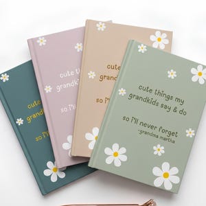 Op de afbeelding: Vier notitieboeken met harde kaft in saliegroen, beige, oudroze en donkerblauwgroen, elk voorzien van witte madeliefjes en de tekst "cute things my grandkids say & do so i'll never forget -grandma martha." Een roségouden pen met een diamant.