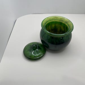 May include: A green jar with a matching lid. The jar is round with a wide opening and a slightly lighter green rim. The lid is flat with a small knob. The jar and lid have a marbled appearance.