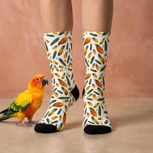 May include: White socks with a repeating pattern of orange, green, and blue leaf shapes. The socks have black heels and toes. A yellow and green parrot with an orange head and beak is standing next to the socks.