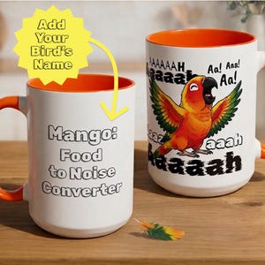 Könnte beinhalten: Zwei weiße Keramikbecher mit orangefarbenen Innenräumen und Griffen. Ein Becher zeigt den Text "Mango: Food to Noise Converter" und einen gelben Stern mit dem Text "Add Your Bird's Name". Der andere Becher hat eine bunte Papageienillustration und die Laute "AAAAAAH" und "Aa! Aaa! Aa!"