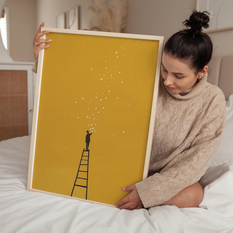 May include: A framed art print featuring a minimalist illustration on a mustard yellow background. The artwork depicts a person on a ladder reaching for stars. The frame is a light wood color.