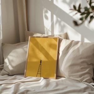 May include: A framed art print featuring a yellow background with a silhouette of a person on a ladder reaching for stars. The artwork is in a light wooden frame and rests on a bed with pillows.