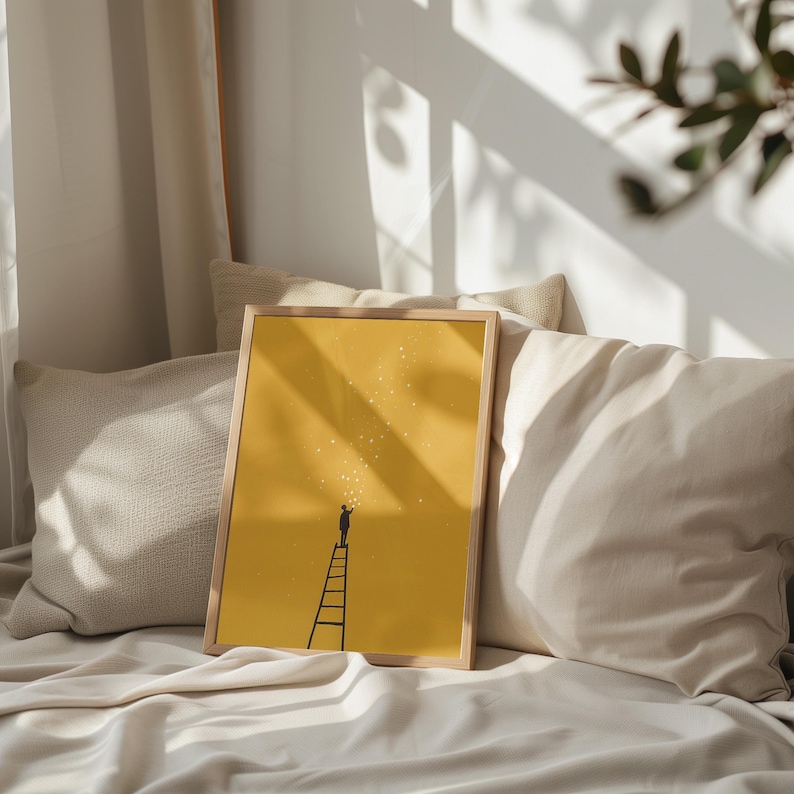May include: A framed art print featuring a yellow background with a silhouette of a person on a ladder reaching for stars. The artwork is in a light wooden frame and rests on a bed with pillows.