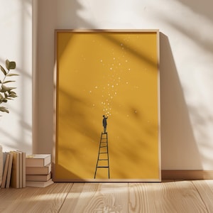 May include: A framed art print showing a silhouette of a person on a ladder reaching for stars. The artwork has a mustard yellow background with white star illustrations. The frame is light brown, and the print leans against a white wall.