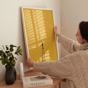 May include: A framed art print featuring a person on a ladder reaching for stars against a yellow background. The artwork is in a white frame and is being held by a person. The print has a minimalist design and is suitable for home decor.