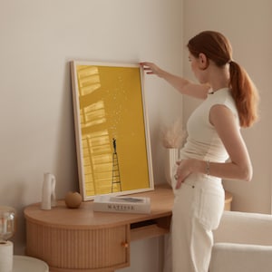 May include: A framed art print featuring a yellow background with a black silhouette of a person on a ladder reaching for stars. The print is in a light wood frame. The word "MATISSE" is on a book on the desk.