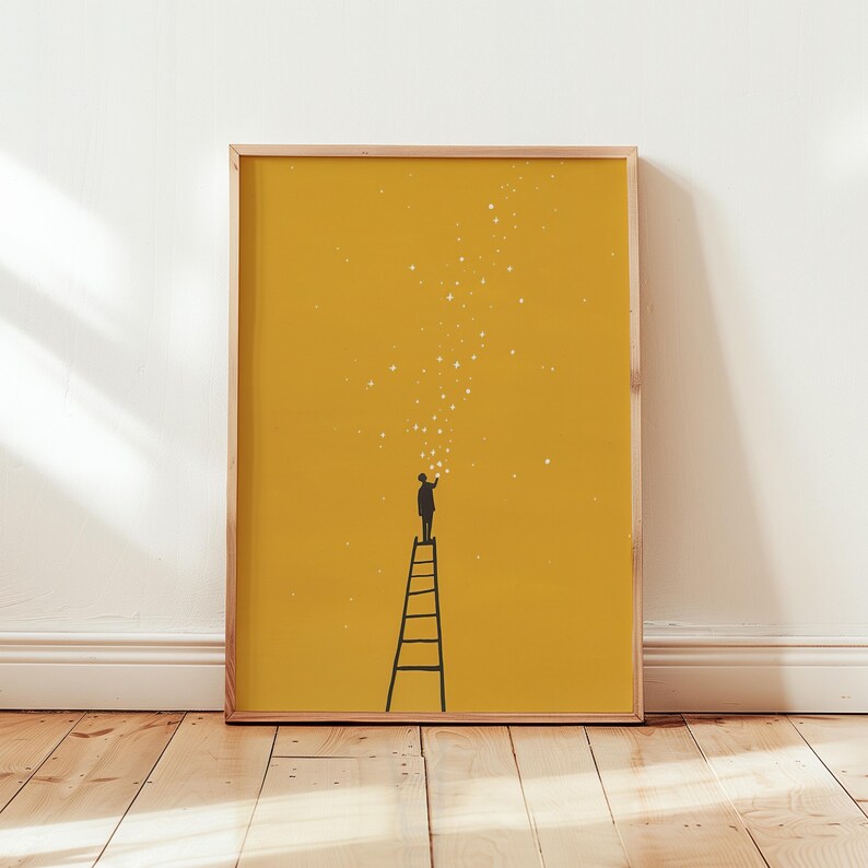 May include: A framed art print featuring a silhouette of a person on a ladder reaching for stars. The background is a solid mustard yellow, and the frame is a light wood color. The artwork is minimalist and inspirational.