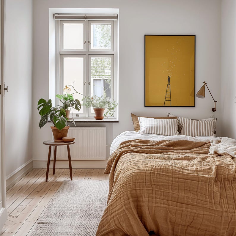 May include: A bedroom scene with a mustard-colored bedspread and striped pillows. A framed yellow poster with a ladder and figure hangs above the bed. A small wooden table holds a potted plant, and a window with plants sits in the background.