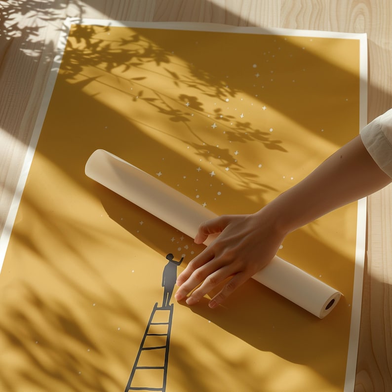 May include: A rolled-up, off-white poster rests on a mustard-yellow print featuring a silhouette of a person on a ladder reaching for stars. The print has a white border and is illuminated by sunlight, casting shadows of leaves.