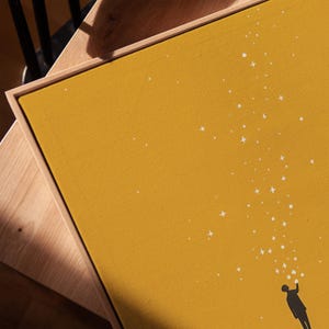May include: Mustard yellow canvas art print featuring a silhouetted figure reaching up towards a stream of white stars. The artwork is framed in a natural wood tone, creating a minimalist and modern aesthetic.