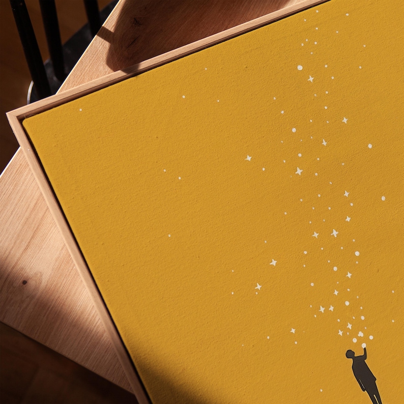 May include: Mustard yellow canvas art print featuring a silhouetted figure reaching up towards a stream of white stars. The artwork is framed in a natural wood tone, creating a minimalist and modern aesthetic.