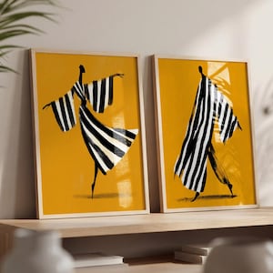 May include: Two framed art prints featuring stylized figures in black and white striped garments against a solid mustard yellow background. The left print shows a figure with arms outstretched, while the right print depicts a figure in a long flowing robe.