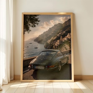 May include: A framed wall art print featuring a dark green vintage Porsche sports car parked on a coastal road. The scene overlooks a calm sea and a hillside town with buildings nestled into the cliffs under a soft, golden sunset sky.