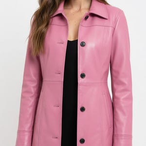 Puede incluir: Un abrigo largo de cuero rosa con cuello y botones negros. El abrigo tiene un corte entallado y cierre de botones. La modelo lleva un top negro debajo del abrigo.