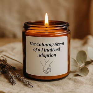 Könnte beinhalten: Eine brennende Kerze in einem braunen Glas. Auf dem weißen Etikett steht "The Calming Scent of a Finalized Adoption" mit einer Zeichnung von Händen. Die Kerze steht auf einer neutralfarbenen Oberfläche. Die Kerze ist etwa 9 cm hoch.