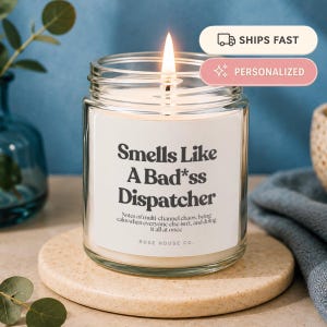 Op de afbeelding: Een kaars in een doorzichtige glazen pot met een brandende lont en een wit etiket met de zwarte tekst Smells Like A Bad*ss Dispatcher, gevolgd door kleinere tekst over multikanaal chaos en kalmte, met de merknaam Rose House Co. onderaan.