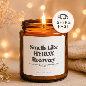 May include: A brown glass jar candle with a white label featuring the black text Smells Like HYROX Recovery, Notes of sore quads and questionable life choices, and ROSE HOUSE CO. The candle has a lit wick with a visible flame and a round sticker.