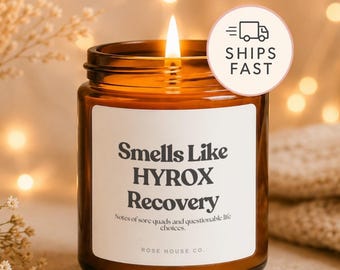 Pode incluir: Uma vela em frasco de vidro castanho com um rótulo branco com o texto preto Smells Like HYROX Recovery, Notes of sore quads and questionable life choices, e ROSE HOUSE CO. A vela tem um pavio aceso com uma chama visível e um autocolante.