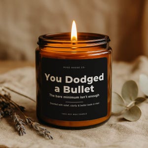 May include: A lit soy wax candle in a brown glass jar. The black label reads "You Dodged a Bullet" with additional text. The candle is on a neutral-toned surface with dried lavender and greenery.