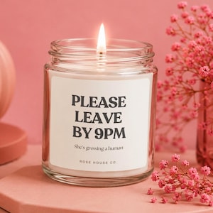 May include: A lit candle in a clear glass jar with a white label. The label reads "PLEASE LEAVE BY 9PM" and "She's growing a human". Pink flowers are in the background. The candle is from ROSE HOUSE CO.