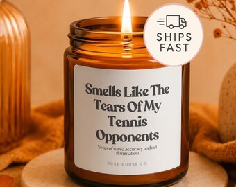 Puede incluir: Una vela encendida en un frasco de vidrio ámbar con una etiqueta blanca que presenta el texto negro Smells Like The Tears Of My Tennis Opponents, Notes of serve accuracy and net domination, y la marca Rose House Co. en el frente.