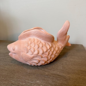May include: A terracotta-colored fish-shaped planter. The fish has detailed scales, fins, and a wide opening on its back for planting. The planter is sitting on a wooden surface.
