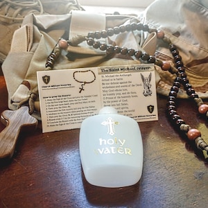May include: An assortment of religious items including a wooden cross, rosary beads, a bottle of holy water, and a card with the Saint Michael Prayer. The bottle has the words "holy water" printed on it.