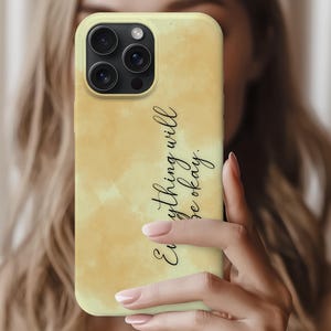 Puede incluir: Una funda de teléfono amarilla con un diseño de acuarela y el texto "Everything will be okay" en escritura negra. La funda tiene un recorte para la cámara y se sostiene frente a una persona.