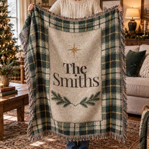 May include: A woven throw blanket featuring a green, cream, and gold plaid pattern with fringed edges. The center panel is cream-colored with the text The Smiths, a gold star icon, and two green pine branch illustrations at the bottom.