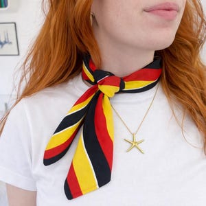 May include: A colorful scarf with black, red, and yellow stripes is tied around the neck. A gold starfish pendant hangs from a delicate chain. The person is wearing a white t-shirt.