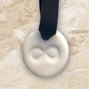 May include: A round, off-white pendant with an embossed infinity symbol, suspended from a black ribbon. The pendant is centered against a beige and brown marble background. The design is simple and elegant.