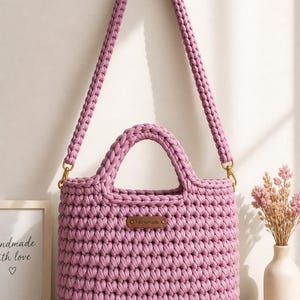 Pode incluir: Bolsa de mão de crochê roxa feita à mão, com alça de ombro e alças superiores. A bolsa tem formato retangular com textura tecida e ferragens douradas. Uma pequena etiqueta de couro marrom diz "Handmade".
