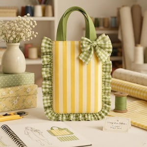 May include: A rectangular tote bag featuring vertical yellow and white stripes, accented with green and white gingham ruffles along the sides and bottom. The bag includes matching green gingham handles and a decorative bow on the upper right corner.