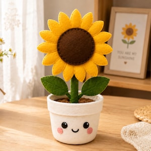 May include: A felt sunflower plant in a white pot with a smiling face. The sunflower has bright yellow petals and a dark brown center. The pot features two black eyes, a small black mouth, and two pink circular cheeks, all crafted from felt.