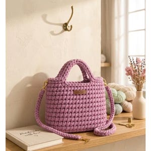 Könnte beinhalten: Eine handgefertigte, lavendelfarbene Häkeltasche mit einem oberen Griff und einem abnehmbaren Schultergurt. Die Tasche hat goldfarbene Hardware und ein kleines, rechteckiges Lederetikett. Ein Buch mit den Worten "Handmade with Love" ist sichtbar.