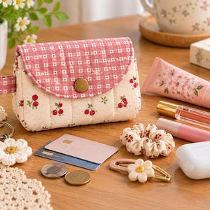 May include: A quilted fabric pouch featuring a pink and white gingham flap with a gold button closure and a cream-colored body patterned with small red cherries. The pouch is accompanied by a key fob, credit cards, coins, a hair scrunchie, a hair clip, a tube of cream, lip products, and a white earbud case.