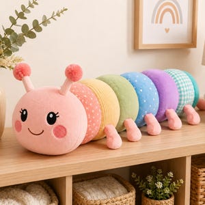 May include: A plush caterpillar toy featuring a round pink head with antennae, large black eyes, and rosy cheeks. Its segmented body consists of various colors and patterns, including polka dots, stripes, and gingham, with small pink feet attached.