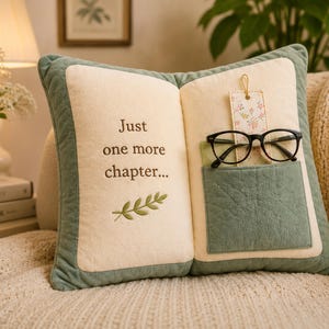 May include: A rectangular throw pillow shaped like an open book with a sage green border and cream-colored pages. The left side features the embroidered text "Just one more chapter..." and a small leaf icon. The right side has a sage green pocket.