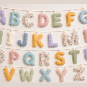 Könnte beinhalten: Eine dekorative Girlande mit den sechsundzwanzig Buchstaben des Alphabets, die jeweils als weiche, gepolsterte Stoffform gestaltet sind. Die Buchstaben hängen an vier weißen Schnüren und zeigen eine Vielzahl von einfarbigen Pastelltönen und Blumenmustern.