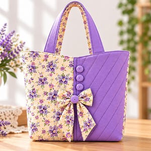 May include: A quilted tote bag featuring a split design with one side in a cream fabric with a purple floral print and the other side in solid purple with diagonal stitching. The bag includes three purple buttons and a matching floral fabric bow.