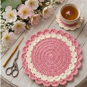 May include: A pink and cream crocheted placemat and matching coaster with a cup of tea and flowers. The placemat is round with a scalloped edge. A crochet hook and scissors are also on the table.